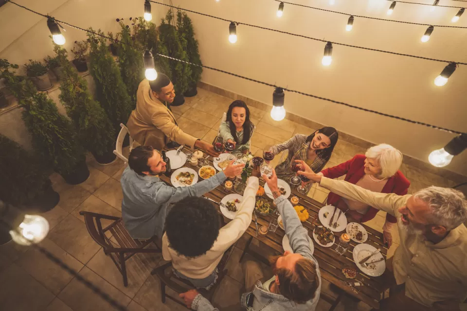 Diverse group of people sitting around a table doing a toast on a patio with warm bistro lights.